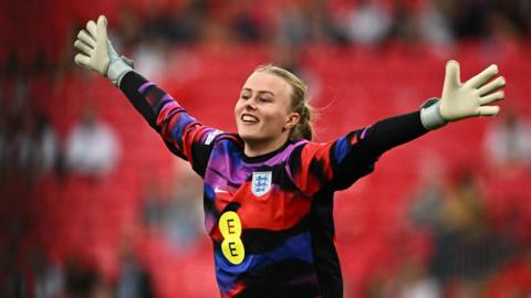 Hannah Hampton in a colourful red, purple, blue and black goalie's jersey, with her hands stretched out wide and a smile on her face. Her blonde hair is tied bak and she is wearing white goalie gloves. Stands are blurred behind her.