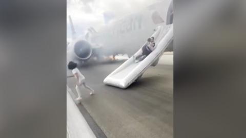 A man slides down an inflatable evacuation slide from a plane as a carrying a child. Another child runs away from the slide. There is smoke in the background, coming from the plane.