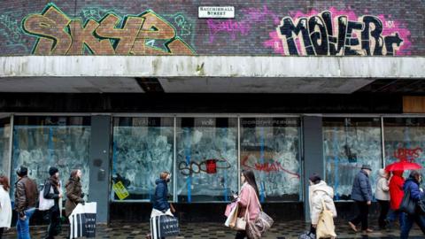 Shoppers walk past a closed shop on Glasgow's Sauchiehall Street. There is graffiti on the windows and on the walls. 