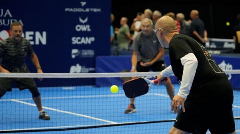 Three men playing pickleball on a blue court, with one player in the foreground about to strike the ball over the net to two opposition players he is facing.