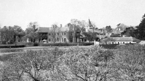 A black and white image of the hospital from the distance. A Victorian looking building can be seen from behind some bushes.