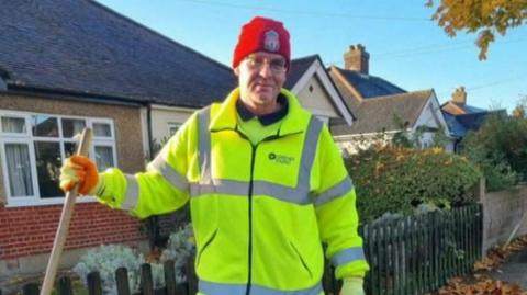 Wayne Broadhurst is pictured wearing high visibility uniform with gloves and a hat while holding a broom stick. Houses are visible in the background.