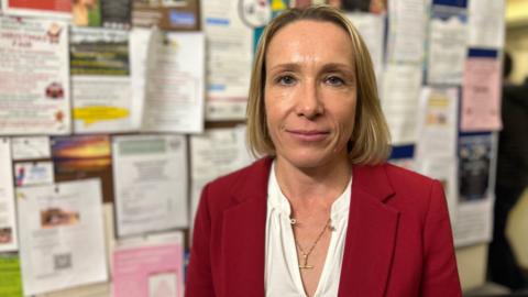 Helen Morgan has a blonde bob and is wearing a white shirt with a red suit blazer. She has a gold necklace on and is standing in front of a board with dozens of posters and leaflets pinned to it.