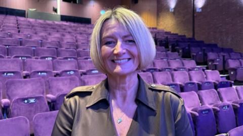 A woman in a theatre with purple lights, standing in front of rows of purple seating. She has a blonde bob and is wearing a black shirt