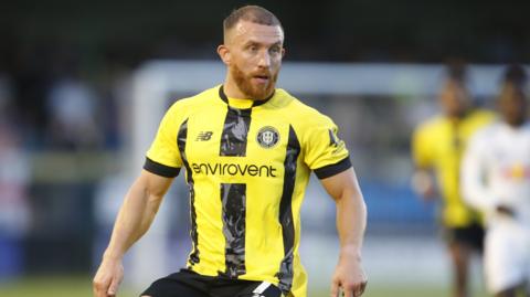 George Thomson intently watching the ball in midfield, wearing home club yellow and black shirt
