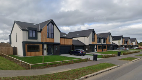 A row of modern detached homes featuring dark grey roofs, dark window frames, wood pannelling and grey and cream cladding. Each has a plain rectangle of grass at the front. The sky is cloudy.