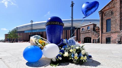 A mooring post painted in royal blue with a bronze ring bearing Michael Jones' name, surrounded by flowers, balloons and an Everton scarf