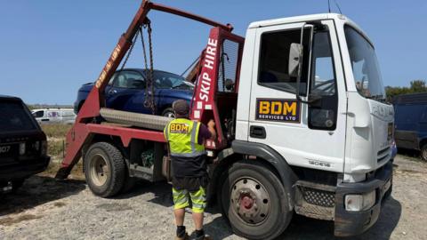 A large white pick-up is seen with a blue car on the back. A man in high-viz clothing is standing beside the vehicle with his hand on what appear to be controls for the back of the truck.