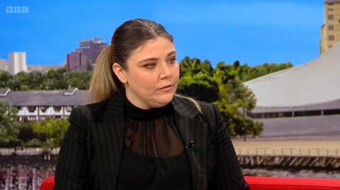 A woman with long brown hair and wearing a black top and black suit jacket is sitting on the red BBC Breakfast sofa. Images of Media City in Salford can be seen behind her.