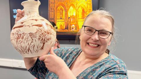 Hilary Steele, who has glasses and tied-back blond hair, looks at the camera and smiles. She is holding up a cream-coloured jug with a decorative red pattern on it.