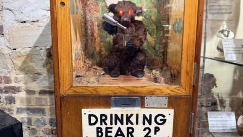 A brown bear toy in a wood and glass case. The bear, which has lit-up red eyes, appears to be pouring contents of a silver bottle into a silver cup. There is a sign under the case that reads 'Drinking Bear 2p'.