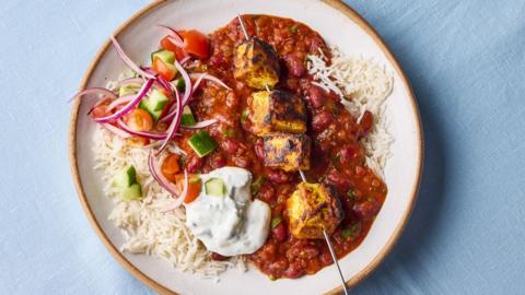A skewer of tofu sits on a red masala sauce and rice, with diced tomato and cucumber and a dollop of yoghurt also on the plate