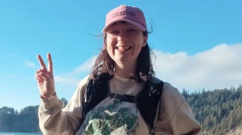 Becky in a pink hat wearing a beige jumper is standing smiling, holding two fingers up in the "peace" sign. A forest can be seen in the background, and blue sky above it. 