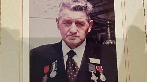 Ben Gimbert has dark wavey hair and is wearing a black blazer with four medals on his lapels, he has a white shirt and a purple spotted tie. Behind him is an old stone wall.