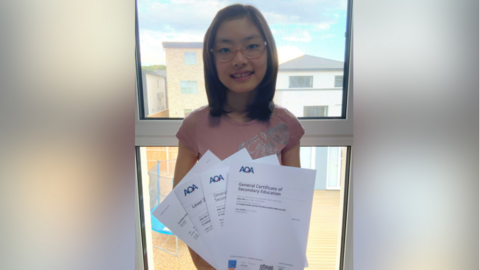 A smiling Ariel with her results. She is holding four sheets of paper. She is in front of a window and buildings are seen behind her. She is wearing glasses.