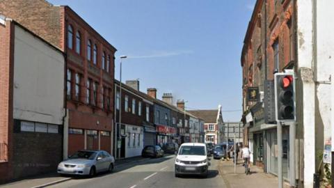 A road with terraced red-brick properties on either side, most of which have shop fronts on the street. A white can is stopped at a red traffic light and a man is riding a bike on the pavement.
