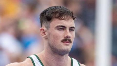 A close-up of Joe Chadwick's head and shoulders as he stands waiting on a start line. He has short dark hair and moustache and wears a white running vest with green piping.