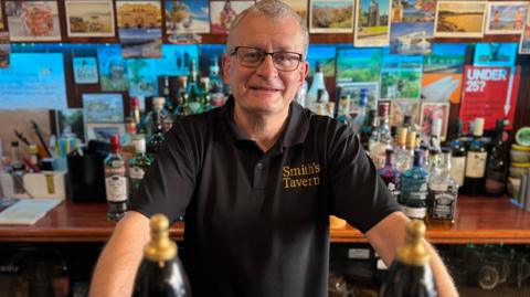 Mark Grist, pub landlord, behind the bar wearing a black t-shirt with a "Smith's Tavern" logo. The wall behind is lined with spirit bottles and posters