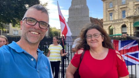 Ed Hill wears a mid-blue polo shirt and black-framed glasses. He is standing next to Angie Nash who is wearing a red top and spectacles, with a bag over her shoulder. They are standing in front of the war memorial in Exeter which has a St Geroge's flag attached to the railings in the centre and a Union flag in the background to the right of the picture.