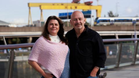 A woman in a pink top and a man in a black shirt. Yellow Harland & Wolff cranes are in the background.