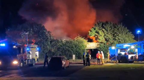 Police officers and firefighters oversee a fire, there are large clouds of orange smoke and two fire engines