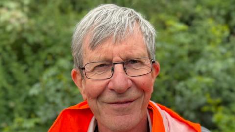 A man wearing an orange high visibility jacket in front of a hedge. He is smiling straight at the camera and wearing glasses.