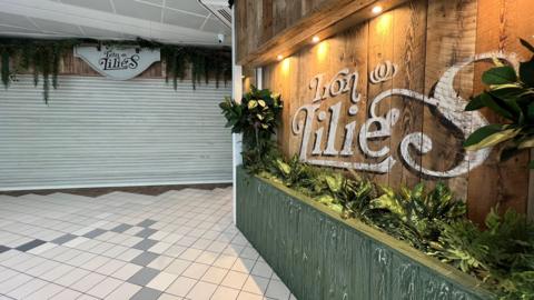 A view of a restaurant and bar inside a shopping centre. White shutters are down over the entrance and there is a wooden side panel to the right of the picture with the words Iron Lilies in white written across it.
