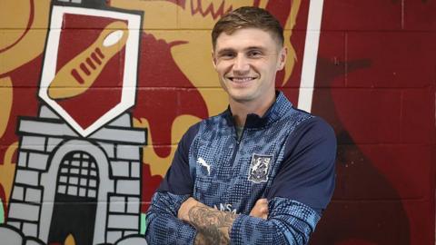 Northampton Saints striker Harvey Saunders folding his arms in front of the club crest on the wall after signing.