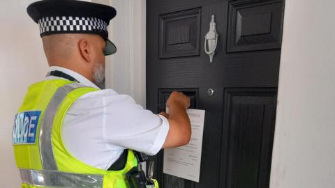 A police officer wearing his police cap, a yellow sleeve-less hi-vis jacket and a white shirt-sleeved shirt pins a closure order on the property's black door.