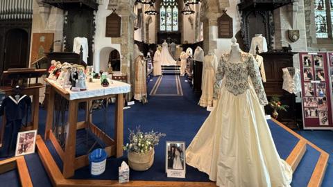 An embroidered wedding dress stands on a mannequin with several others in the background. The dress is a centrepiece in the middle of St Andrew's Church with other wedding-themed items on display in an exhibition. 