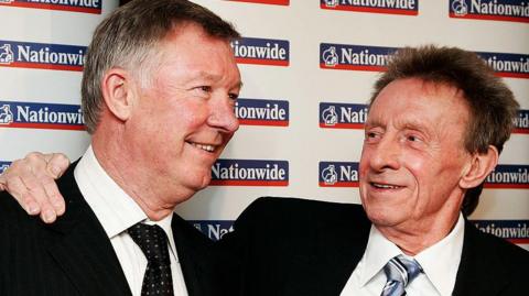 Denis Law, wearing a black suit, white shirt, and blue and grey striped tie, stands with his arm around Sir Alex Ferguson, wearing a black suit, white striped shirt and black tie