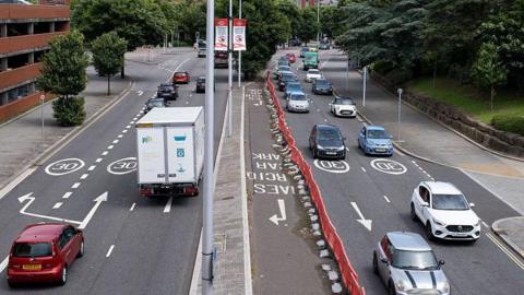A dual carriageway with plenty of cars and a white van driving in both directions. There are also roadworks on one side. 