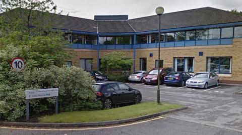 General street view image of the 2 The Pavilions in Ashton. A two-storey building, sandstone on the ground floor and with blue framed glass on the top floor and a grey roof. There is a car park to the front with greenery
