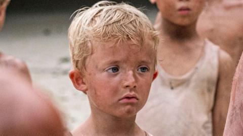 Freddie Lee-Grey in a promotional shot from the Lord of the Flies series. He has dirt on his face and his blond hair is dishevelled. In the background but cropped out there are other children in grubby clothes.