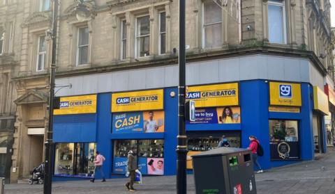 Cash Generator pawn shop with blue and yellow signage in a grand Grade II-listed building in Bradford city centre on the corner of Kirkgate and Bank Street, including pedestrians passing and a bin in the foreground.