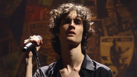 Sombr in a half-unbuttoned black shirt holding a microphone stand and looking moody on stage at the 2025 MTV Video Music Awards