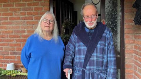 Jan and Graham stand side by side on their doorstep, looking into the camera. Jan, on the left, is wearing a bright blue v-neck sweater, and has long light grey hair and dark-framed glasses. Graham is wearing a checked blue dressing gown with a blue top, and has short dark grey hair and a white beard. He's also wearing dark-framed glasses, and is leaning on a stick.