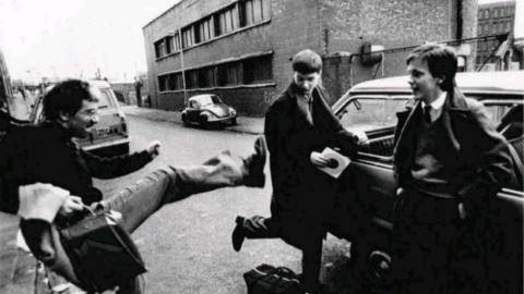 Grainy black-and-white photo of a young Bernard Sumner kicking his leg towards a young Ian Curtis. Rob Gretton is nearby, smiling and leaning against a car. They are standing in a street in front of a brick warehouse.
