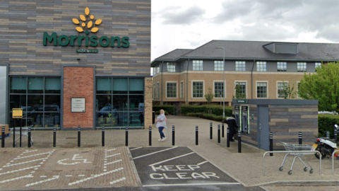 A Google Street view image of a Morrisons which the building finishes half way on the image, and is on the left. On the right is a small building with two ATM machines. A person is standing at one of the machines.