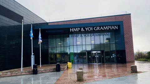 Brick and glass building with sign wording saying 'HMP & YOI Grampian', and flagpoles at the side.