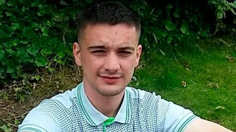 A man with short, dark hair, shaved at the sides, squints at the camera. He is wearing a white polo shirt with green and blue dots. He appears to be sitting on a grass verge and foliage is visible in the background.