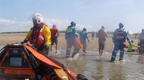 The casualties walking from the lifeboat to the beach, accompanied by lifeboat crew in yellow jackets and blue all-in-one suits