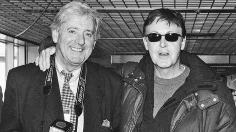 Paul McCartney wearing a jacket and sunglasses has an arm around Dennis Stone, who wears a camera around his neck, at Heathrow Airport