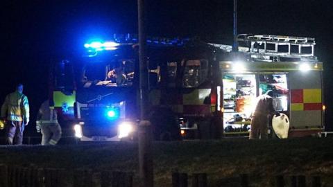 A fire engine with its lights on parked by the side of a grass verge at night. There are five firefighters around the vehicle.