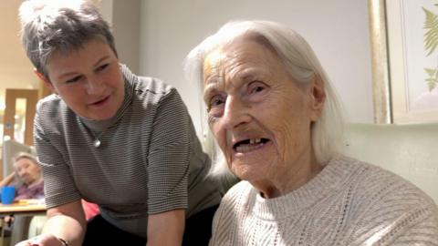 Two women, one with shoulder length grey hair and a white knitted jumper and another leaning over her with a grey top and short grey hair
