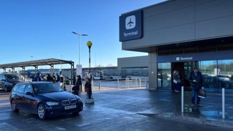 Bristol Airport drop-off parking, with a car in the foreground and people queuing up behind. Signs for the terminal can be seen at the top