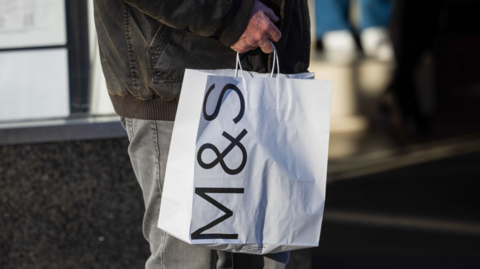 Man in dark winter jacket and grey jeans carries white paper bag with M&S logo on the side 