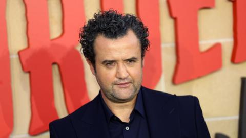 Daniel Mays is at the premier of The Thursday Murder Club in London.  The title of the film is written behind him.  He has olive skin and is wearing a dark blue coloured blazer and shirt.