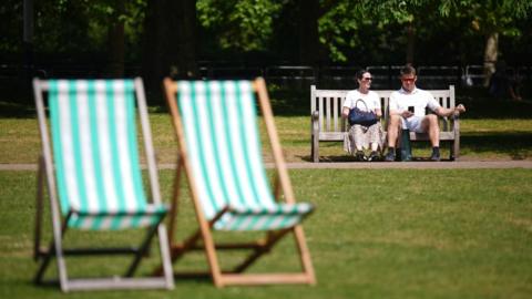 A man and a woman sitting on a bench in a London park on the hottest day of the year so far