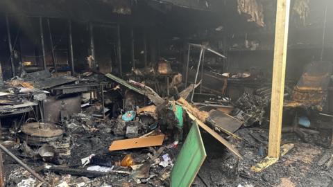An office building completely gutted and destroyed by fire. There are charred, blackened and broken pieces of furniture and items strewn around, with the roof exposed and blown out windows